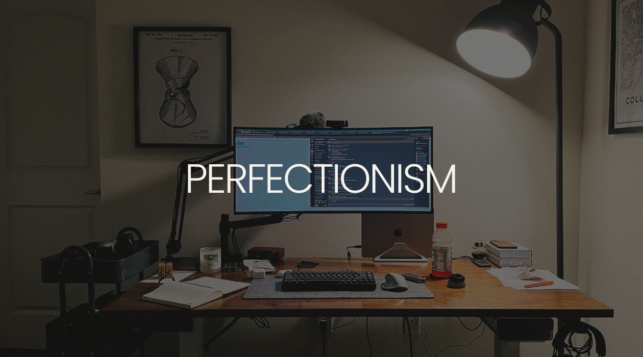Desk with an ultrawide monitor and clutter on it. A graphic of the word Perfectionism is overlaid.